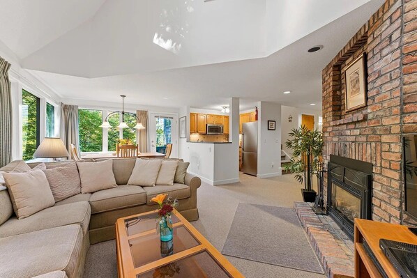 A modern living room with a sectional sofa, a glass coffee table, a fireplace, and a kitchen area visible through an open door.