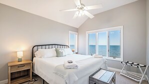 Un dormitorio con cama, mesita de noche, lámpara de noche y vista al mar a través de amplias ventanas.