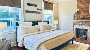 A bedroom with a large bed, a ceiling fan, a fireplace, and a view of the outdoors.