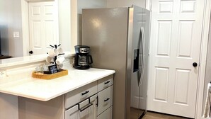 A kitchen with white cabinets, a stainless steel refrigerator, a coffee maker, and a basket of mugs on the counter. - Cielo’s Retreat, A Charming 3-bedroom farmhouse in McKinneyprincenton (McKinney)