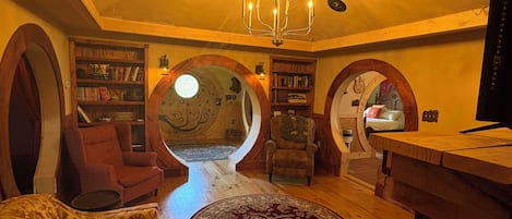 A cozy living room with a wooden floor, a round rug, and a large chandelier. There are bookshelves and a television on the right side.