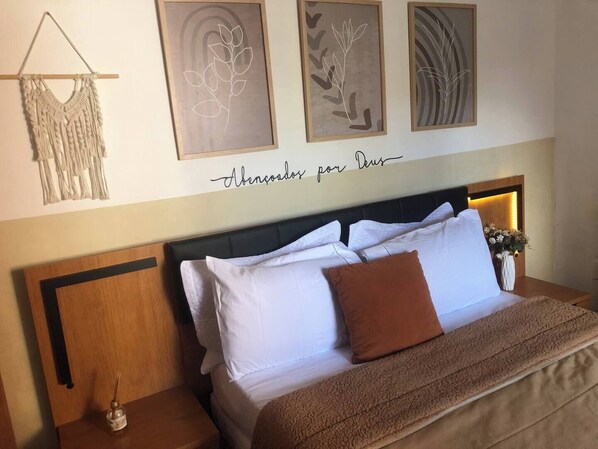 A neatly made bed with white and brown pillows, a macrame wall hanging, and three framed artworks above it.