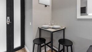 A small dining area with a table set for two, two black stools, a white wall-mounted mirror, and a door with a black frame. - Waou Essence IV Antón Martín - Corta Duración (Madrid)