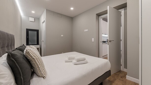 A neatly made bed with white linens and two towels, a grey headboard, and a small bedside table with a lamp. - Waou Essence IV Antón Martín - Corta Duración (Madrid)