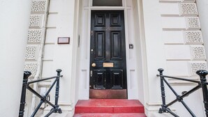 A black door with a number plate, white wall with decorative panels, and black iron railings on red steps. - Blueground | Notting Hill, w/d, nr Hyde Park (London)