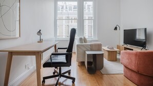 A modern living room with a wooden desk, black office chair, a red sofa, a TV, and a large window with a view of buildings. - Blueground | Notting Hill, w/d, nr Hyde Park (London)