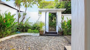 Un sendero de jardín que conduce a una estructura blanca con una puerta, rodeada de diversas plantas y un terreno de grava.