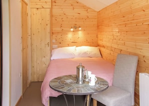 A wooden cabin room with a bed, a small table with a coffee pot, and a chair.
