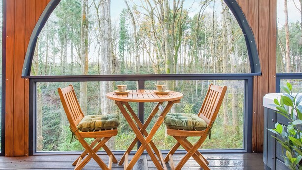 Eine Holzterrasse mit Tisch und zwei Stühlen, ein großes Rundbogenfenster und Blick auf einen Wald.