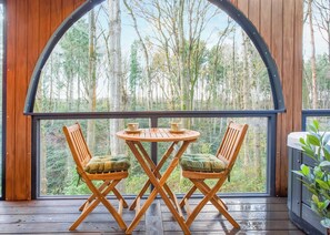A wooden deck with a table and two chairs, a large arched window, and a view of a forest.
