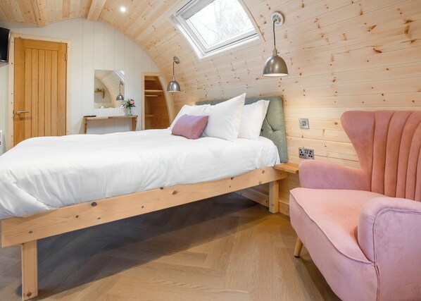 A bedroom with a wooden ceiling, a bed, a pink chair, and a skylight.