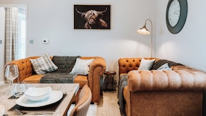 Un salon moderne avec deux canapés en cuir marron, une table à manger avec un plateau blanc et un plateau en verre, et une grande horloge au mur.