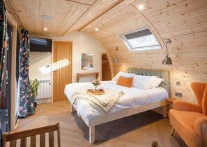 Quarto aconchegante com teto de madeira, claraboia, cama com bandeja de frutas, uma cadeira e mesa de cabeceira.