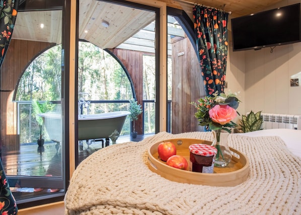 Une pièce confortable avec une grande fenêtre, un plancher en bois, un lit, une télévision et un plateau avec des fruits et un pot de confiture.