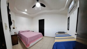 A room with two beds, one with a pink bedspread and the other with a blue and yellow striped bedspread. There is a ceiling fan, an air conditioning unit, and a door in the room. - La Regional (Loreto)