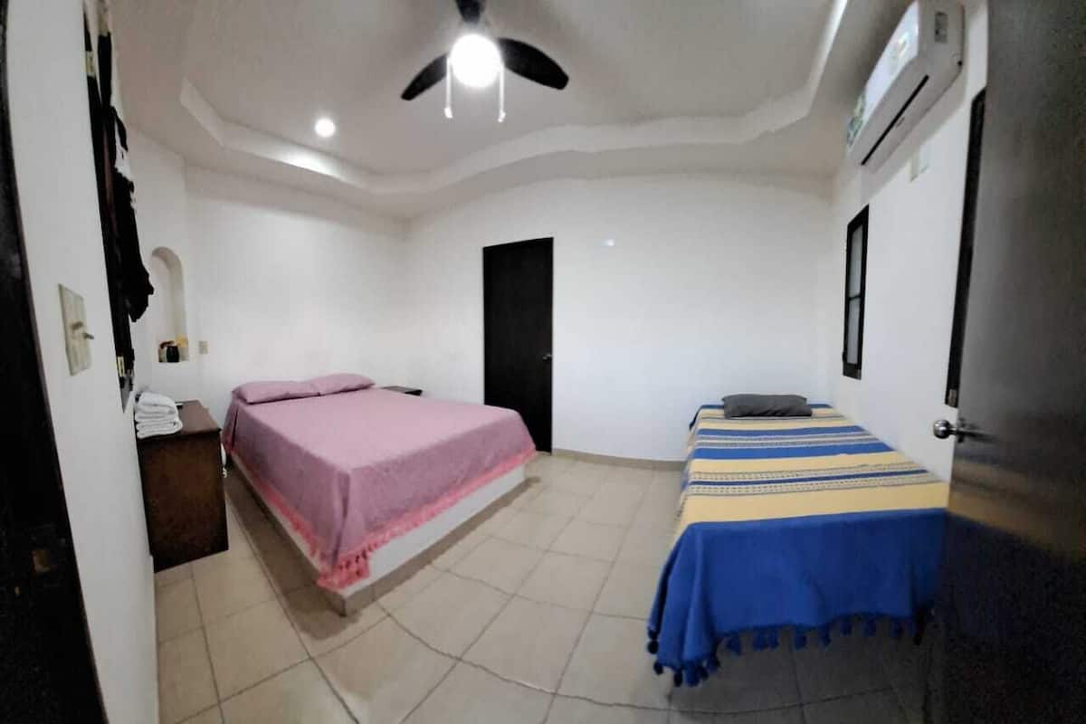 A room with two beds, one with a pink bedspread and the other with a blue and yellow striped bedspread. There is a ceiling fan, an air conditioning unit, and a door in the room.