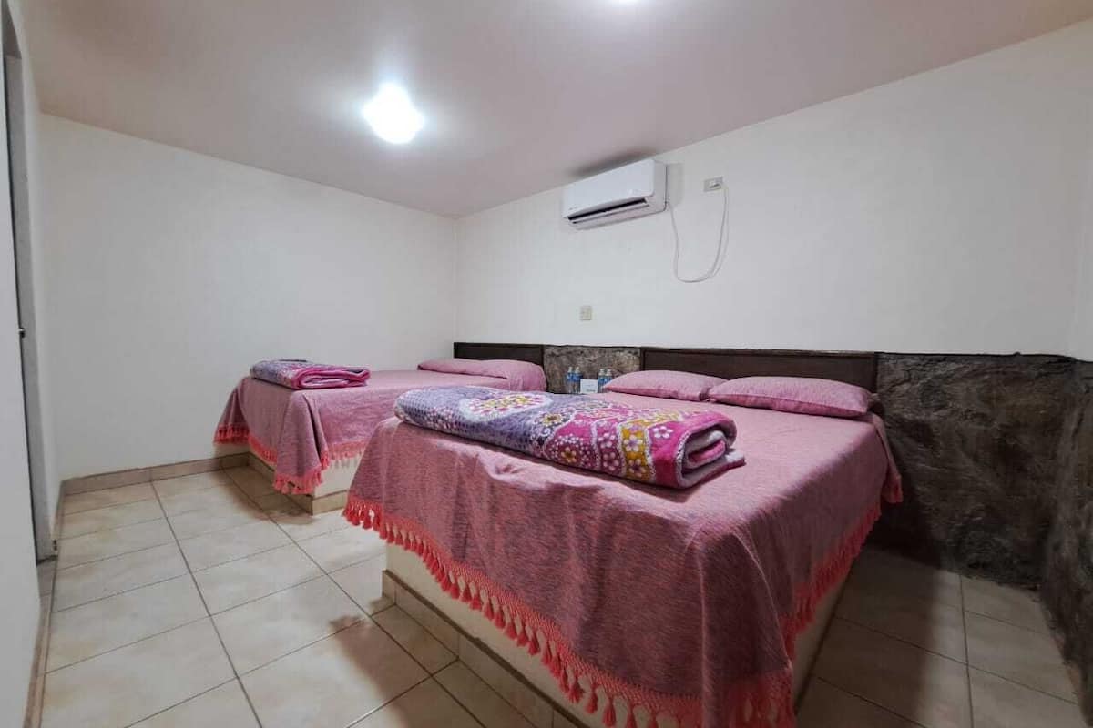 A room with two beds, each covered with pink blankets and a patterned quilt. There is an air conditioning unit on the wall.