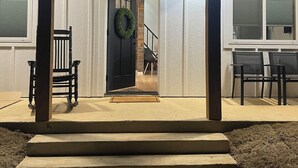 A covered porch with steps leading to a door, a black chair, and a table with chairs.