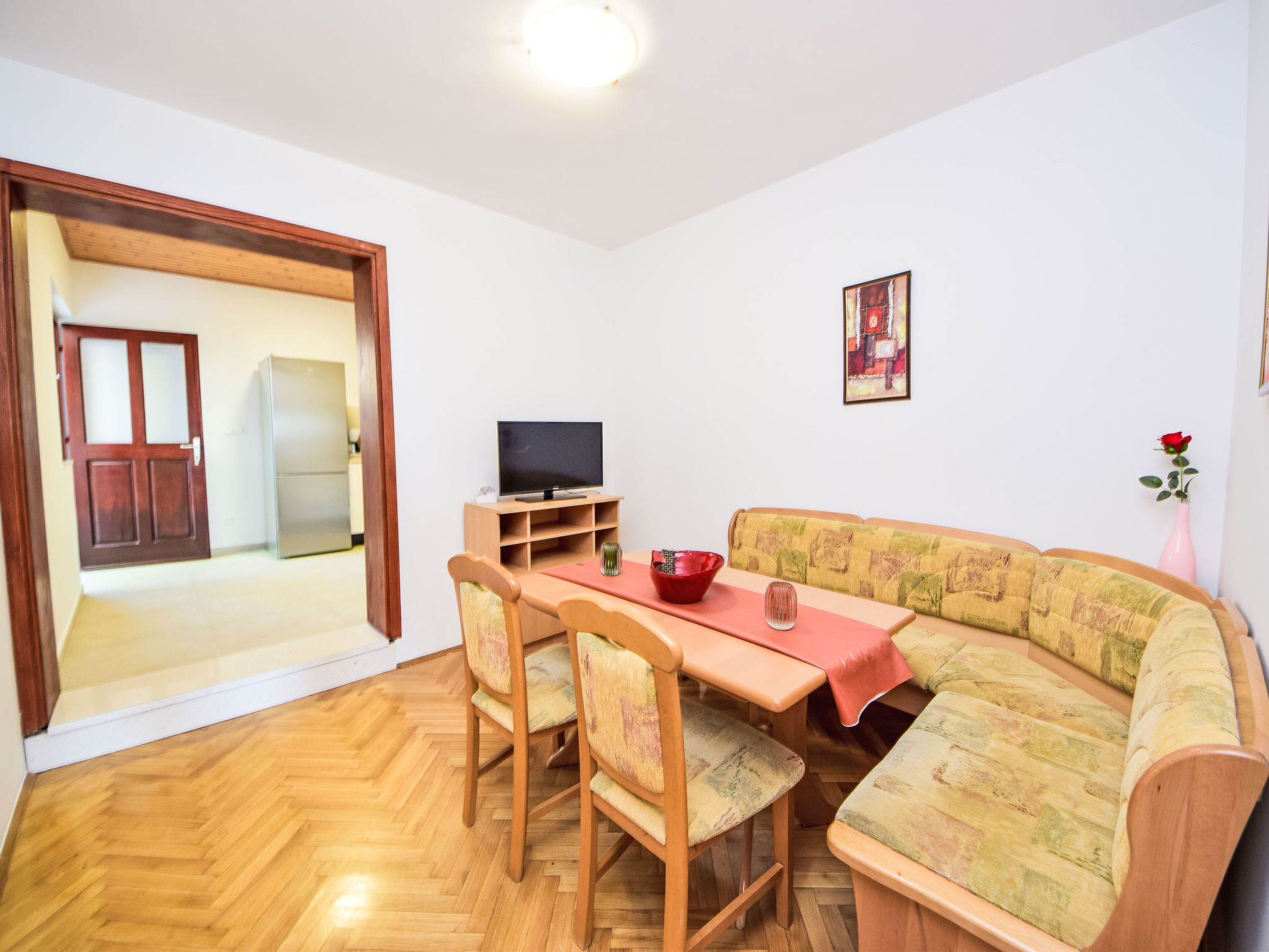 Ein Wohnzimmer mit Holzdielen im Fischgrätmuster, einer Couch, einem Esstisch mit Stühlen, einem Fernseher auf einem Schrank und einer Tür zu einem anderen Raum.
