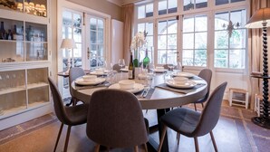 A dining room with a round table set for six, surrounded by six chairs. There is a chandelier above the table, and large windows with curtains on the right side. - Chante le Coq by Interhome (De Haan)