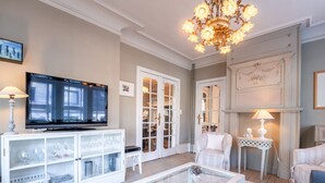 A living room with a television, a glass coffee table, a white armchair, a small side table, and a chandelier. - Chante le Coq by Interhome (De Haan)