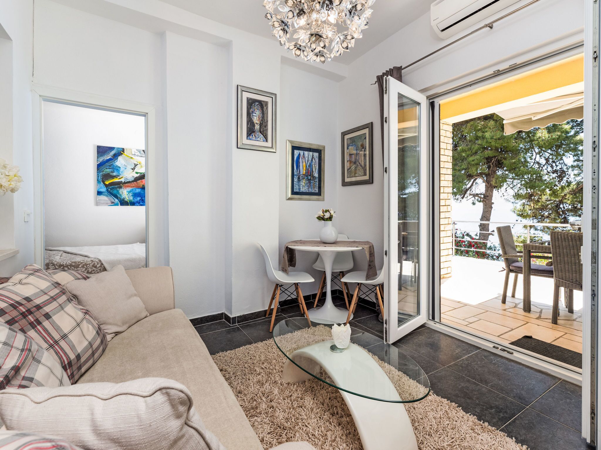 A modern living room with a sofa, a glass coffee table, and a dining area with chairs. There are paintings on the wall and a view of the outdoors through a glass door.