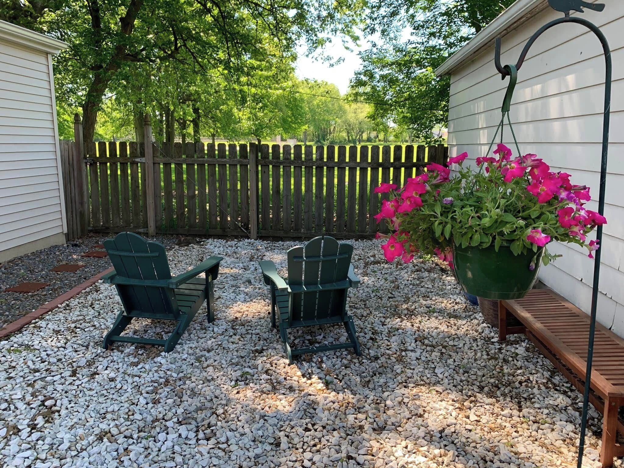 A backyard with two Adirondack chairs, a wooden bench, and a hanging flower pot with pink flowers.