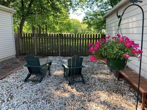 Ein Garten mit zwei Adirondack-Stühlen, einer Holzbank und einem hängenden Blumentopf mit rosa Blüten.