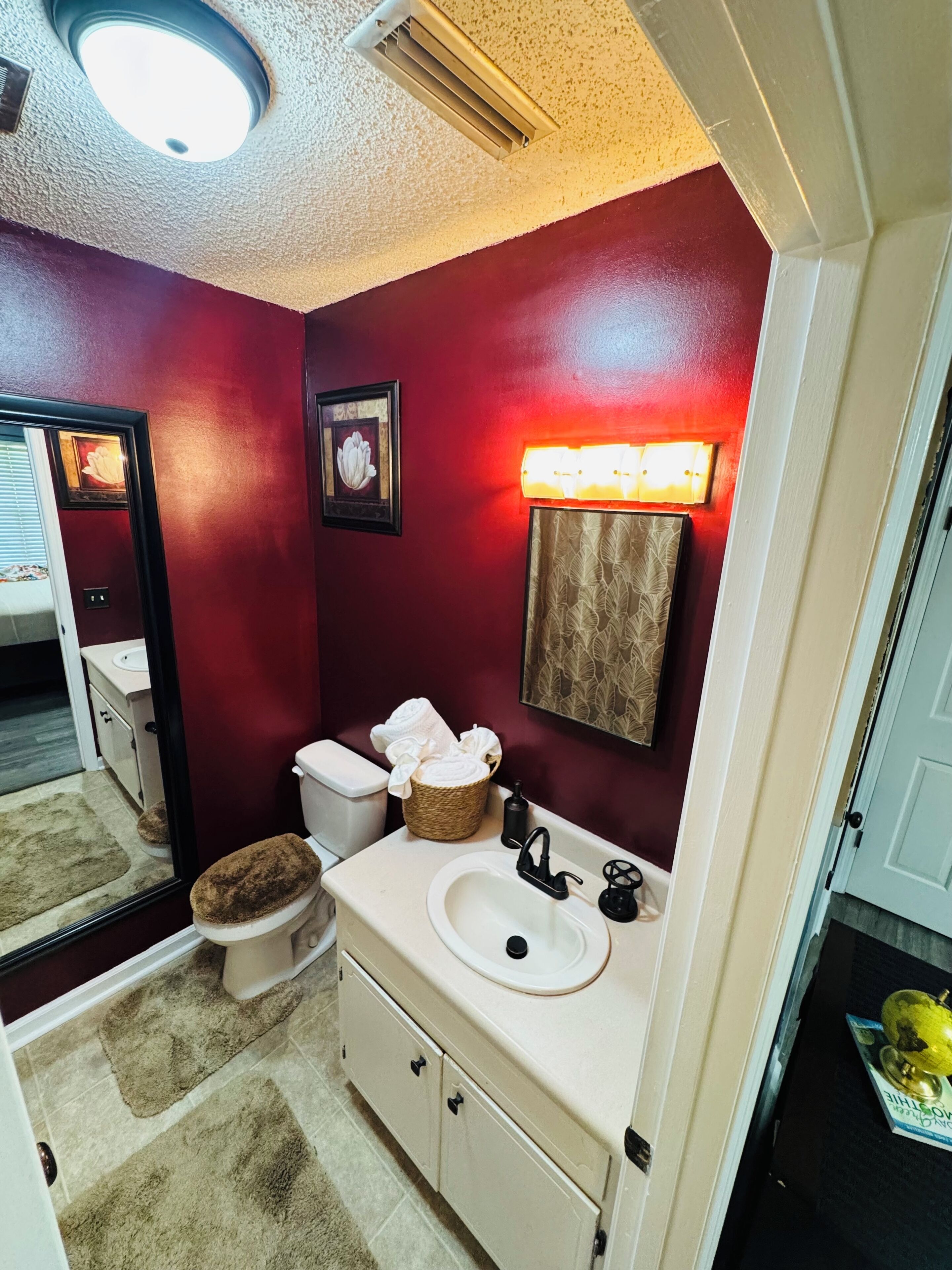 A bathroom with a red wall, a white sink, a toilet with a brown seat, a mirror, and a light fixture above the mirror.
