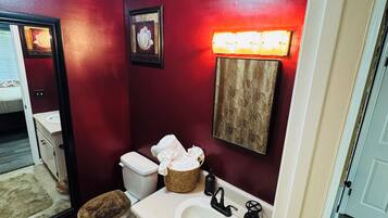 A bathroom with a red wall, a white sink, a toilet with a brown seat, a mirror, and a light fixture above the mirror.