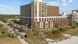 A multi-story hotel with a central courtyard, surrounded by palm trees and a sandy beach.