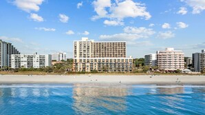 A beachfront hotel with multiple floors, a sandy beach in the foreground, and a clear blue sky.