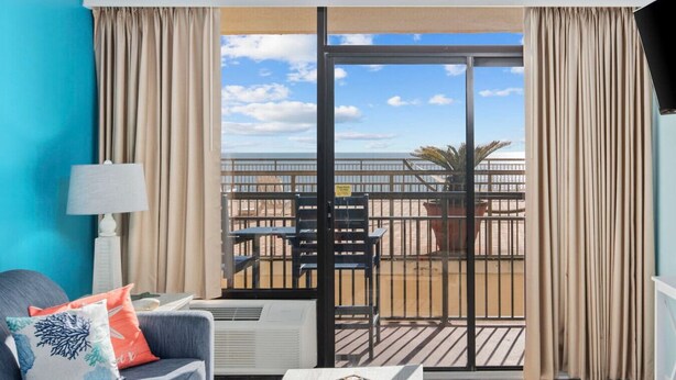 A balcony with a view of the beach, a chair, a table, and a lamp.