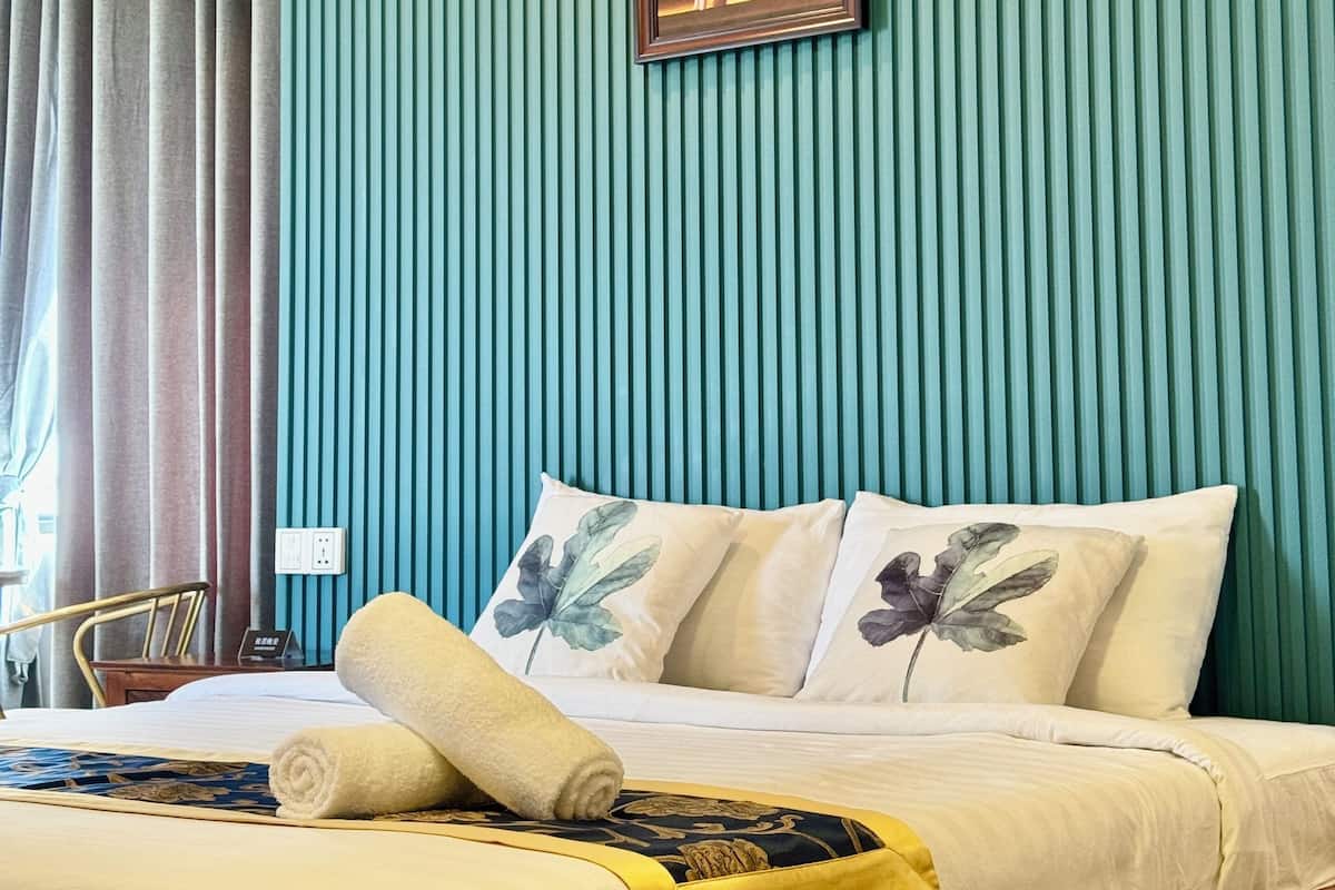 Een hotelkamer met een bed, kussens, een opgerolde handdoek, een schilderij aan de muur en een raam met gordijnen.