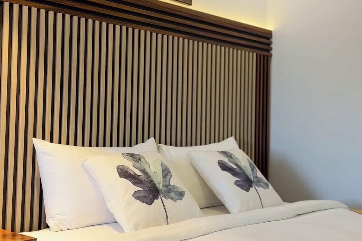Een net opgemaakt bed met witte lakens en sierkussens met bloemenprint, een houten spijbelzijkussen en een schilderij van bloemen aan de muur.