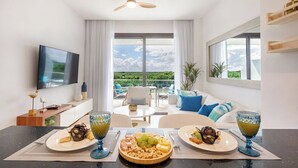 Ein modernes Wohnzimmer mit Essbereich, großen Fenstern, einem Flachbildfernseher und einem Balkon mit Blick auf Himmel und Wolken.