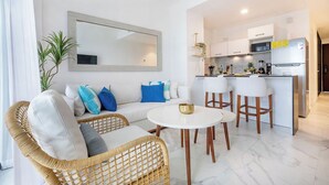 Ein modernes Wohnzimmer mit einer Couch, Sesseln und einem Esstisch. Im Hintergrund ist eine Küche mit weißen Schränken und einem Kühlschrank zu sehen.