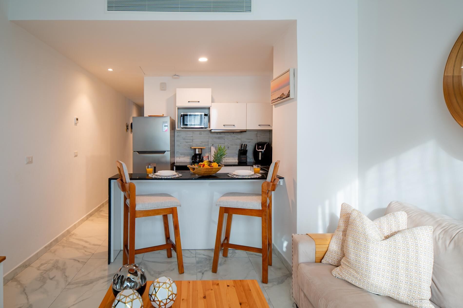 Un espacio habitable compacto con kitchenette, dos sillas de madera, una mesa de centro de madera y un sofá con cojines estampados.