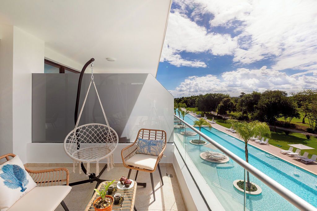A balcony with a hanging chair, a cushioned chair, a small table with plants, and a pool view.