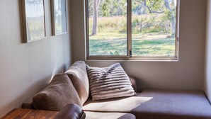 Un rincón acogedor con un banco acolchado y almohadones, una mesa de madera y un ventanal con vistas a un espacio exterior cubierto de césped.