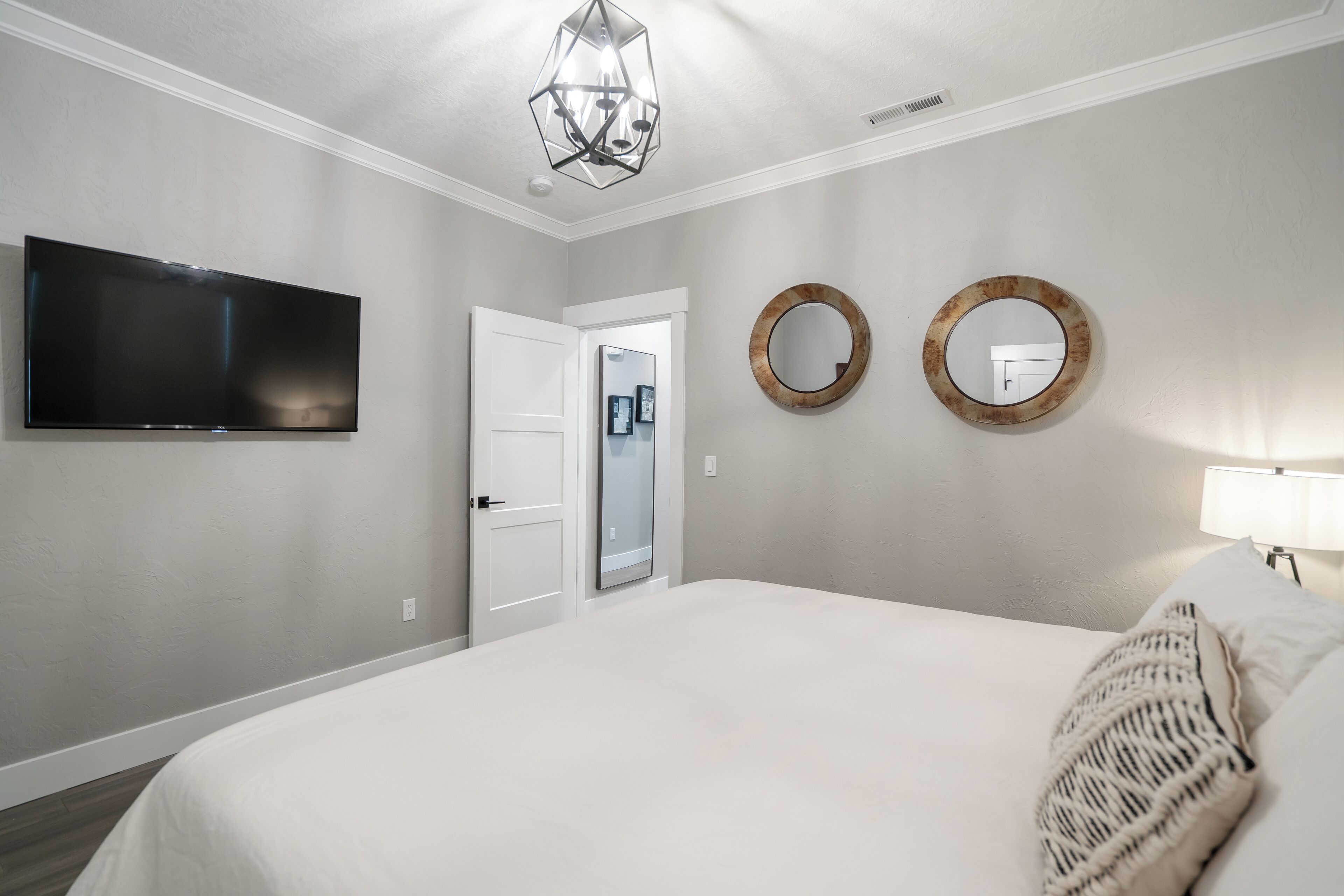 A bedroom with a large bed, a flat-screen TV, two wall-mounted mirrors, and a modern chandelier.
