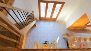 A high-ceilinged room with a wooden staircase, a large window, and a kitchen area with a table and chairs.