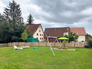 A grassy backyard with a swing set, a wooden fence, and a house with a red roof.
