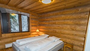 Une chambre dans un chalet en rondins, avec un lit, un petit tabouret en bois et une fenêtre donnant sur des arbres couverts de neige.