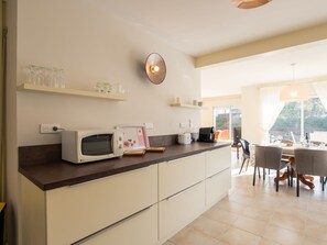 A modern kitchen with a microwave, toaster, and coffee maker on the counter. There are shelves above the counter holding glasses and a teapot. The kitchen leads to a dining area with a table and chairs.