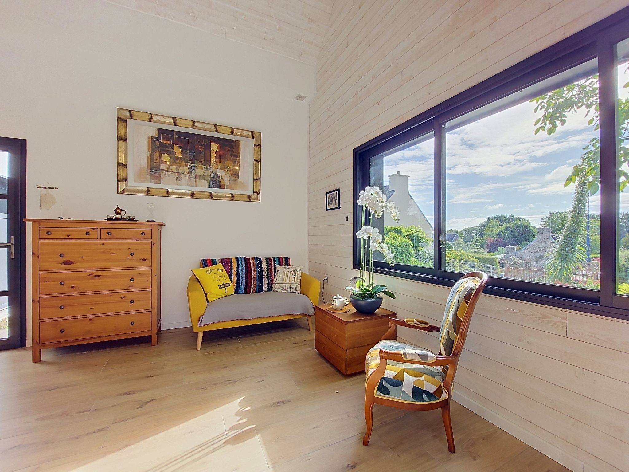 Una habitación con piso de madera, un sofá amarillo, un armario de madera, una silla con cojín estampado, una ventana con vista a follaje y un cuadro en la pared.