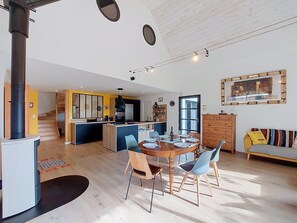 A modern kitchen with a central island, a dining area with a round table and chairs, a staircase leading to another level, and a living area with a sofa and a large window.