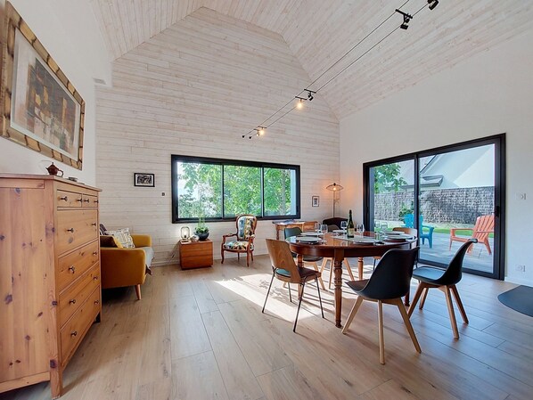 A spacious room with a wooden floor, a dining table set for four, a sofa, a cabinet, and large windows offering a view of the outdoors.