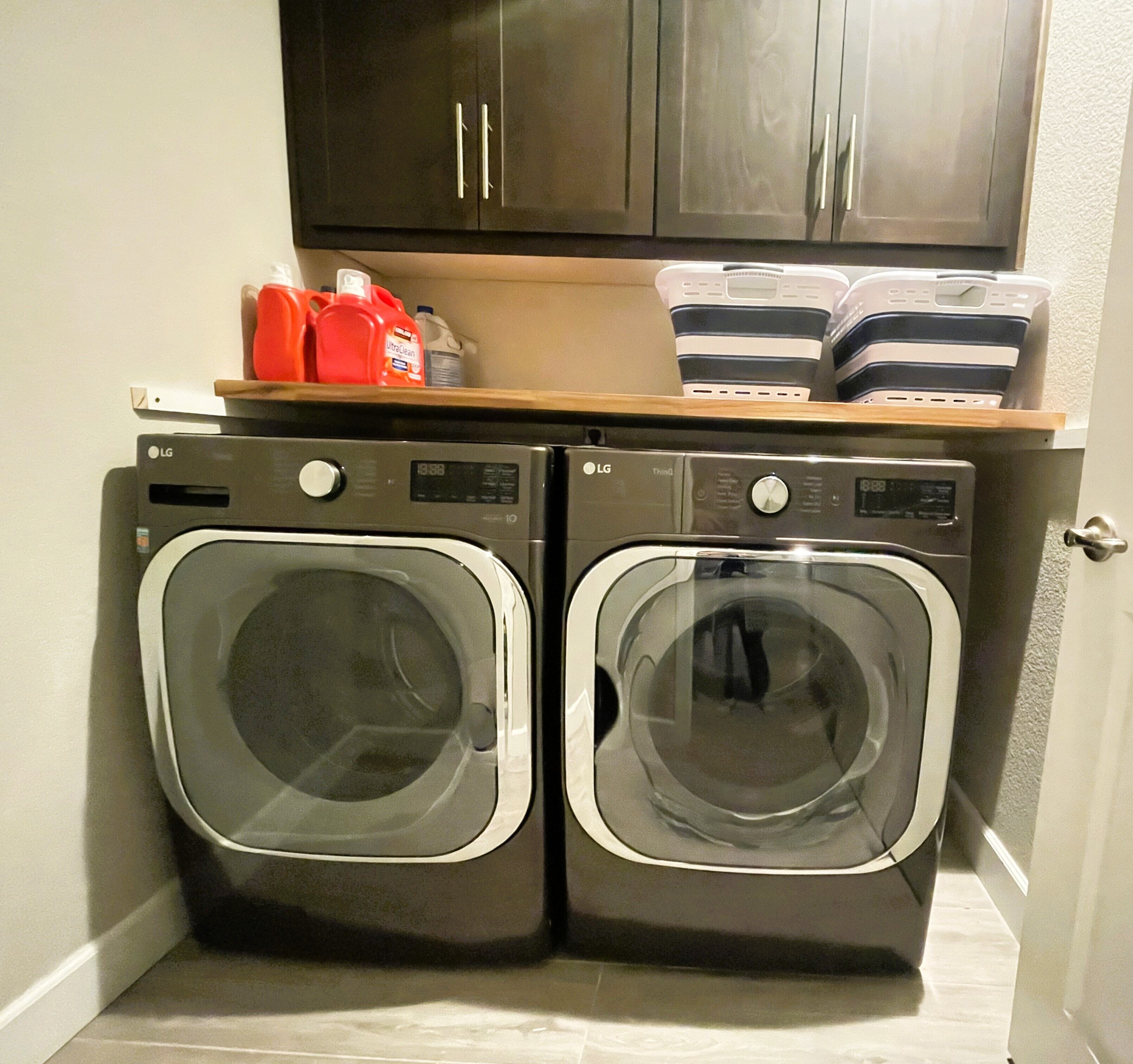 A laundry room with two front-loading washers and dryers, a wooden shelf above with cleaning supplies, and a dark cabinet above.