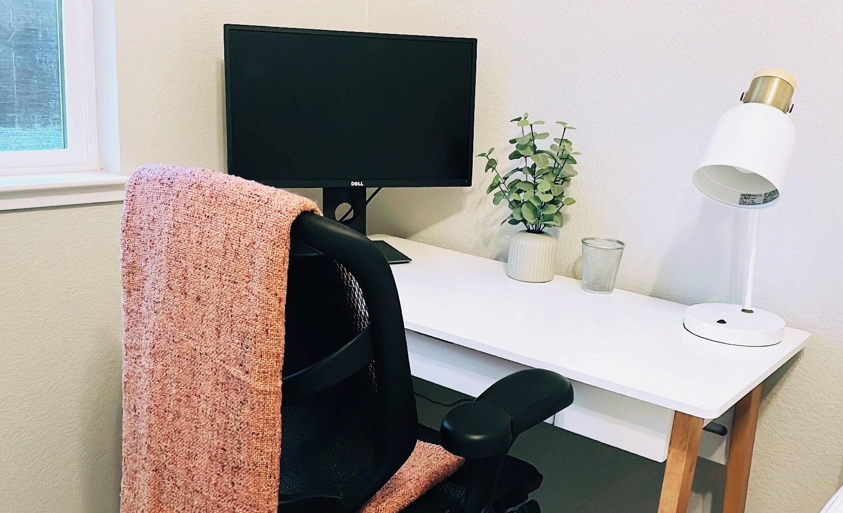 Een moderne kantoorruimte met een wit bureau, een zwart beeldscherm, een groene plant, een witte lamp en een zwarte bureaustoel.